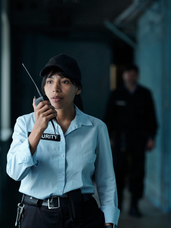 Young female security guard in uniform and cap speaking on walkie-talkie to her colleague while moving along corridor of building